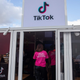 A large glass panneled building with the Tiktok logo on a sign above a door. A person stands in from of the door with a shirt that says "ask me anything about TikTok.